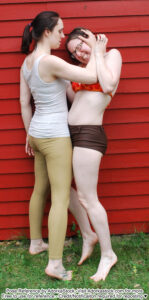 Two white, female pose reference model in standing pose with on holding the others head who seems to be in pain
