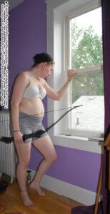 thin, white, female pose reference model in standing pose with a bow in one hand, the other on a window edge looking out