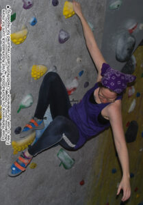thin, white, female pose reference model in hanging pose on a a rock wall, one hand reaching down