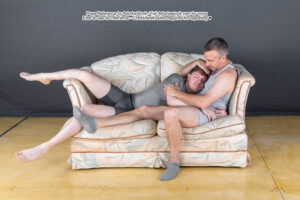 Two men cuddle on a couch, facing right. One man is sitting up and leaning against the arm rest while the other man is lying on top of him, resting his head on the first man's chest.