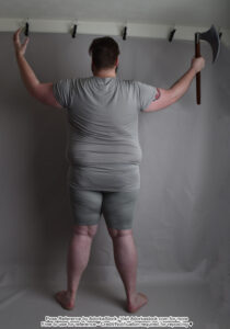 A back facing fat white male model standing with feet shoulder width apart, arms raised with one hand open and the other holding a single blade ax.