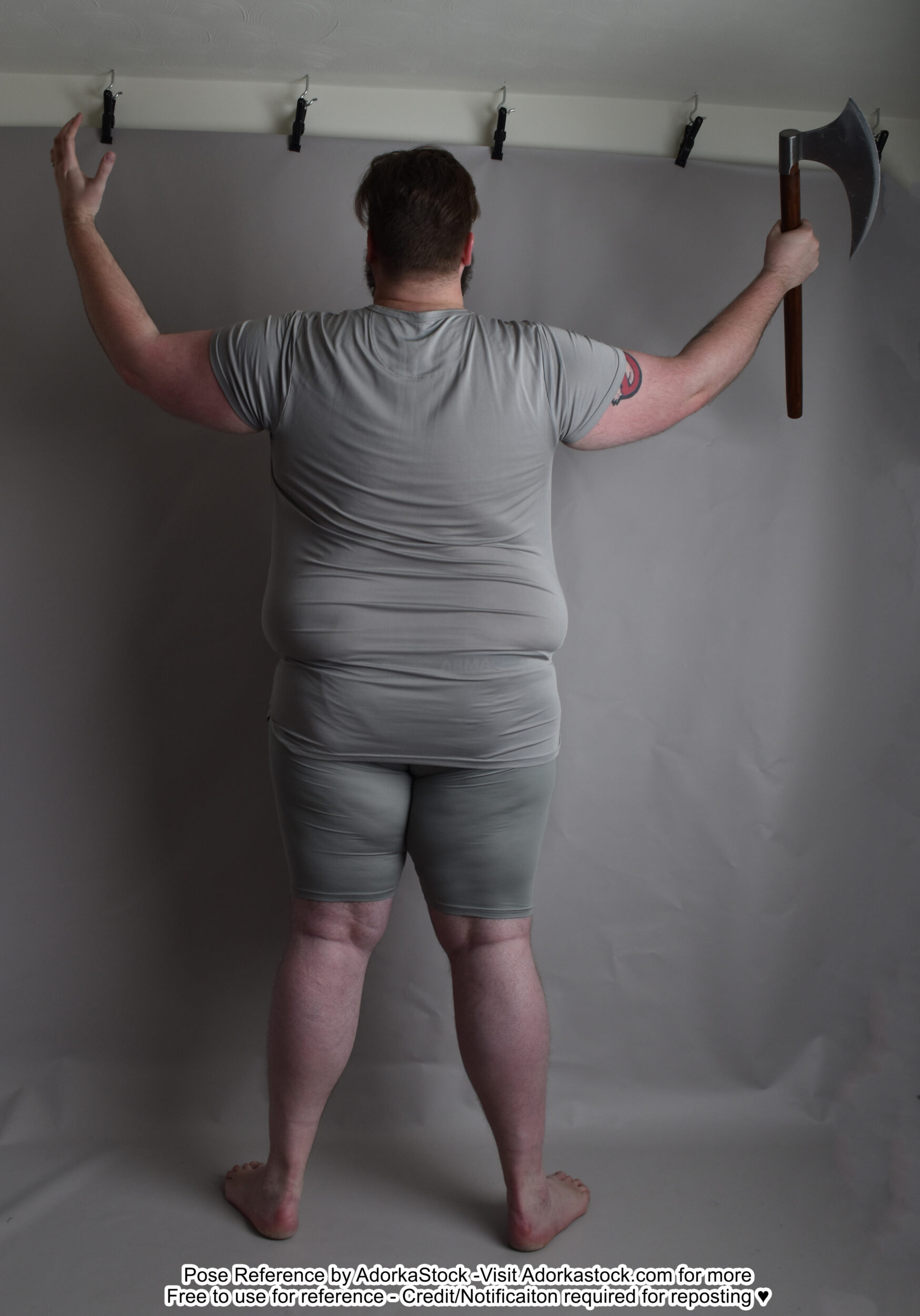 A back facing fat white male model standing with feet shoulder width apart, arms raised with one hand open and the other holding a single blade ax.
