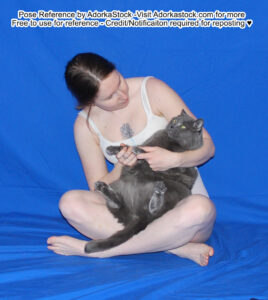 Thin, white, female model is sitting, cross-legged on the ground, holding and looking fondly at a plump and slightly perturbed grey cat in her arms.