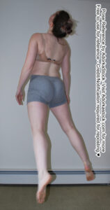 Thin female model with her back to the viewer floats just off the ground. She is looking up, with her arms at her side and her legs apart, toes pointed.