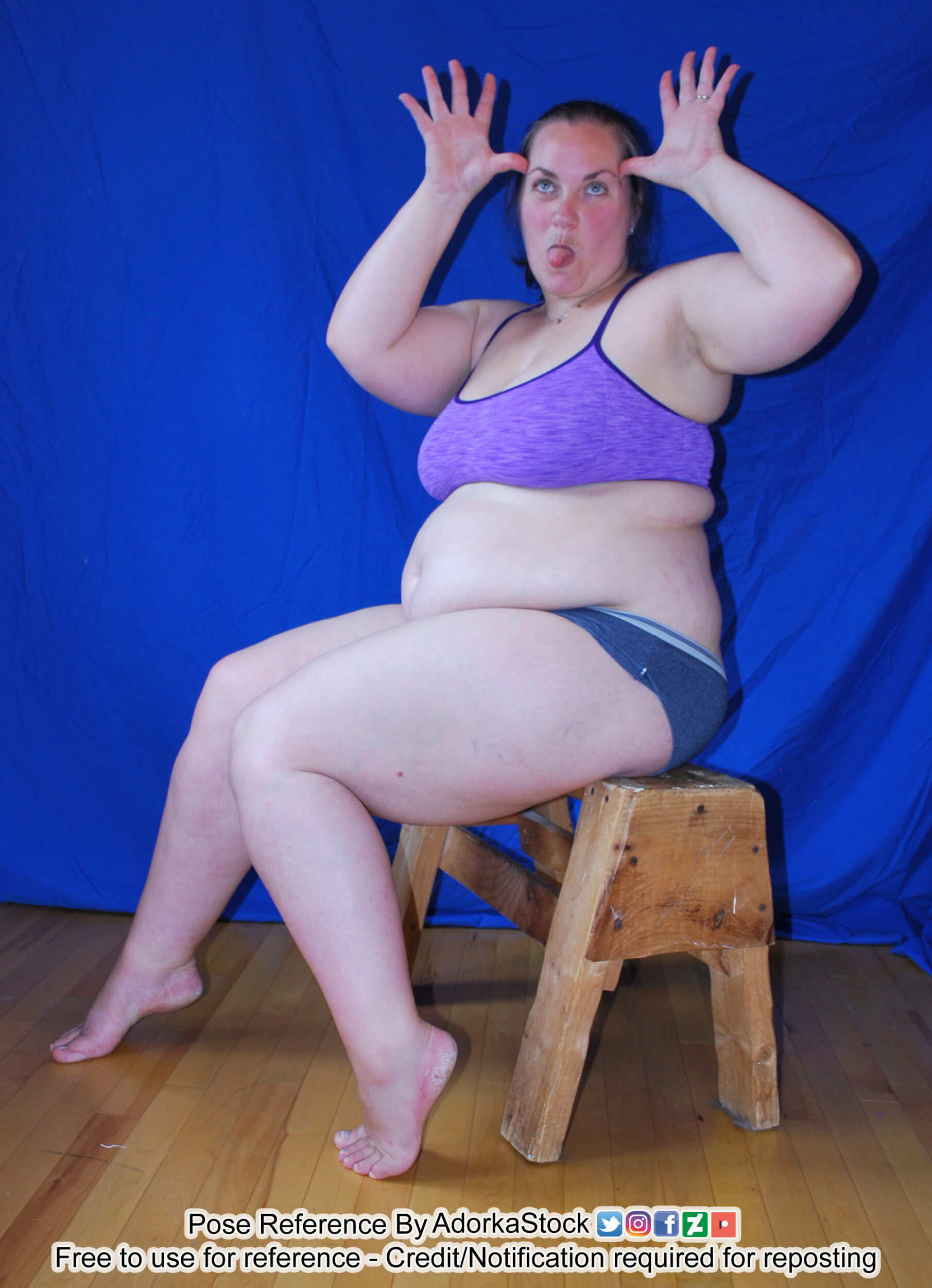 Plus size female model sitting on a stool, making a silly face with eyes crossed and her tongue sticking out. Her hands are up by her head with her thumbs resting on her temples in a comical gesture.