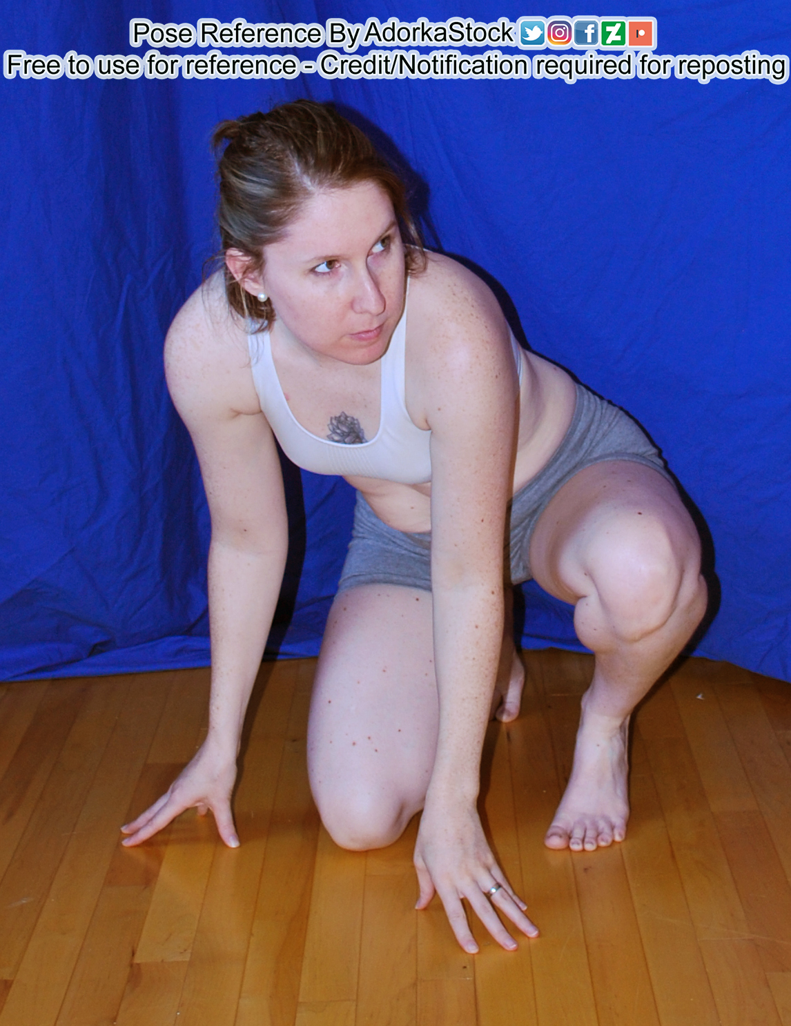 Thin, white, female model is crouching on the floor with one knee down, the other raised. She has both hands on the floor with fingertips touching and she's looking off to the right with some amount of tension.