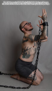 thin white male model kneeling with chains on his wrists which he's raising towards the sky, looking up