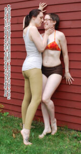 Two thin, white female models one taller than the other, holding the shorter one up against a wall by her neck