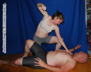 Thin white female model over a larger male model laying on the floor, she is about to punch him in the face