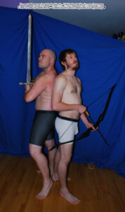 two male models standing back to back one looking serious with a sword the other holding a bow looking tired