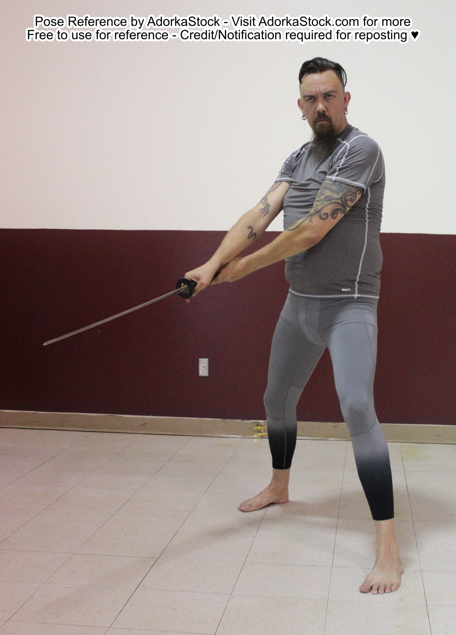 An athletic white male model dressed in form-fitting clothes stands at a three-quarter angle with a wide stance while holding a sword with both hands The blade is pointing slightly downward and out to the left; he is looking forward.