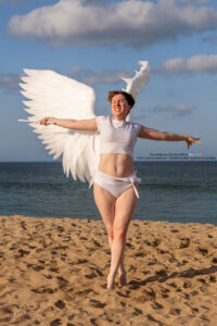 thin white female model wearing huge wings with arms out to the side one holding a wand as if casting magic