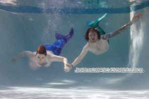 thin white female model and thin white male model swimming underwater holding hands wearing mermaid tails