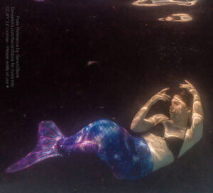 A white woman wearing a multicolored mermaid tail in dark black water, her "tail" bent up towards her.