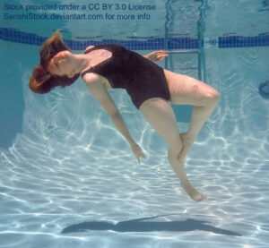 A white woman wearing a black one piece bathing suit floating underwater, arms extended, chest upward, one leg bent up.