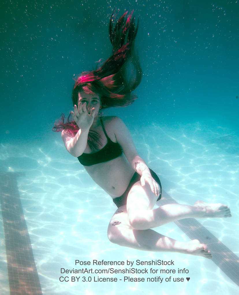 A white woman in a two piece black bathing suit underwater, hair extending up towards the surface, legs bent up, one hand outstretched towards the viewer like she is waving.