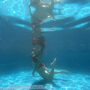 A woman in a two piece black bathing suit, head touching the surface of the water, one knee touching the bottom of a pool, appearing to sink.