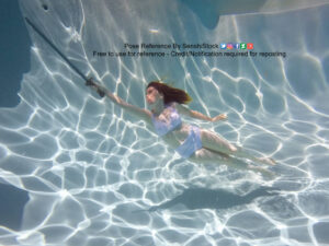 White woman wearing a lavender two piece bathing suit, holding a sword by its hilt and pointing it up as she swims through the water upward.