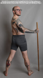 thin male model standing facing mostly away but looking over his shoulder while holding the top of a staff that is touching the ground