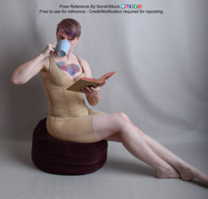 Model sitting on a cushion reading a book in one hand while sipping a cup with the other