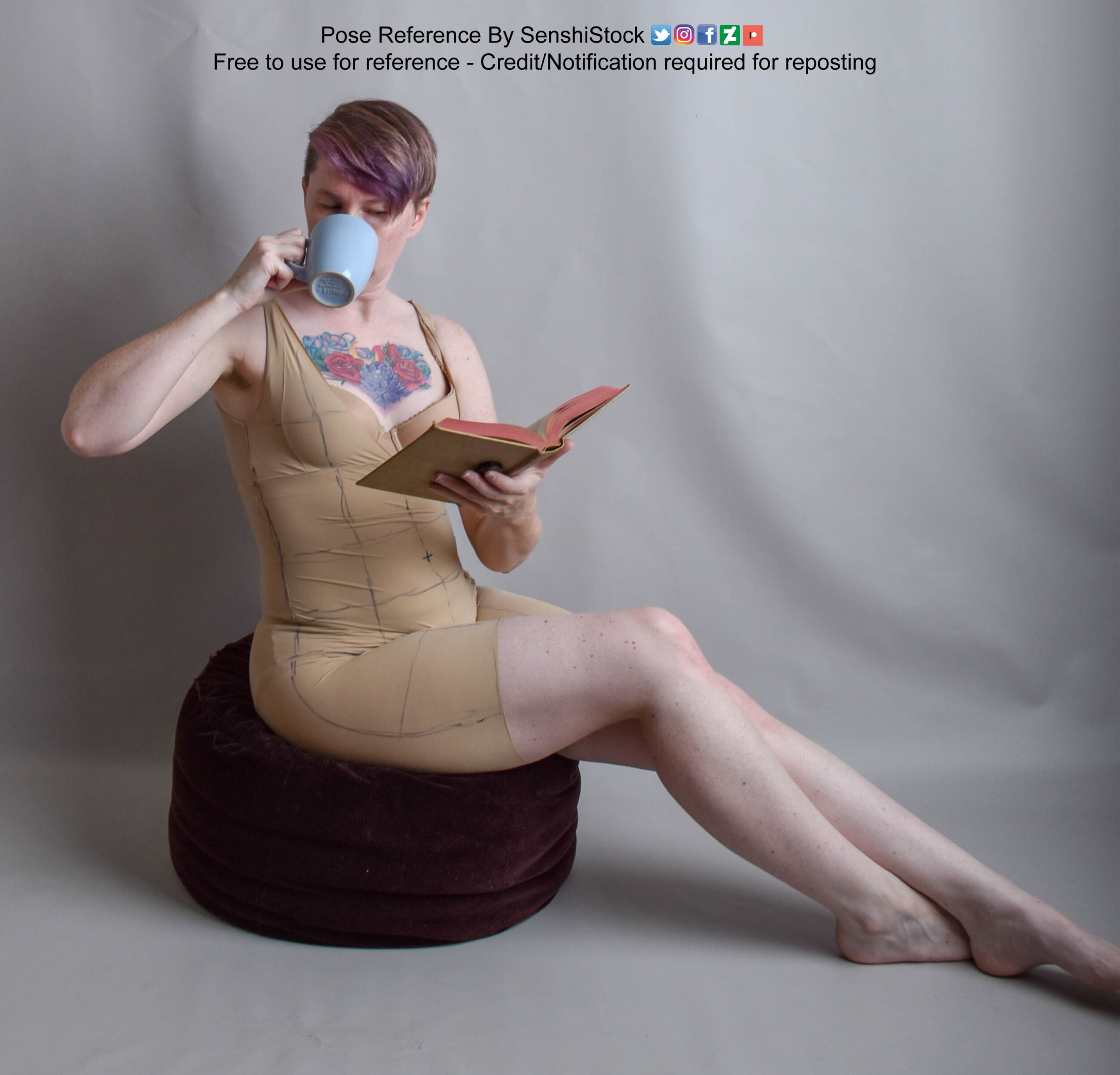 Model sitting on a cushion reading a book in one hand while sipping a cup with the other