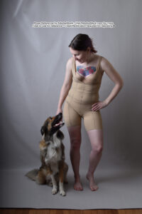 thin white female model standing with her hand on a hip reaching down to pet a fluffy English Shepherd sitting on the floor beside her