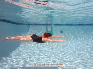 thin white female model swimming underwater horizontally, arms outstretched as if flying
