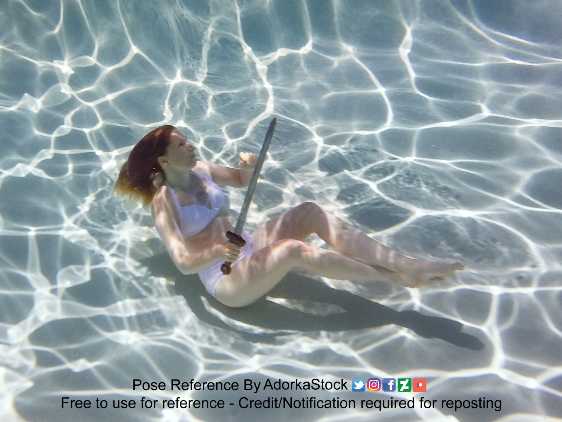 thin white female model underwater with a sword in hand facing the surface