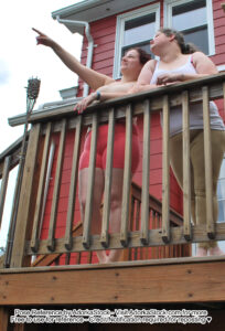 Two models are standing along a deck rail, one is pointing up the other is leaning and it's from a ground perspective looking up at the models.